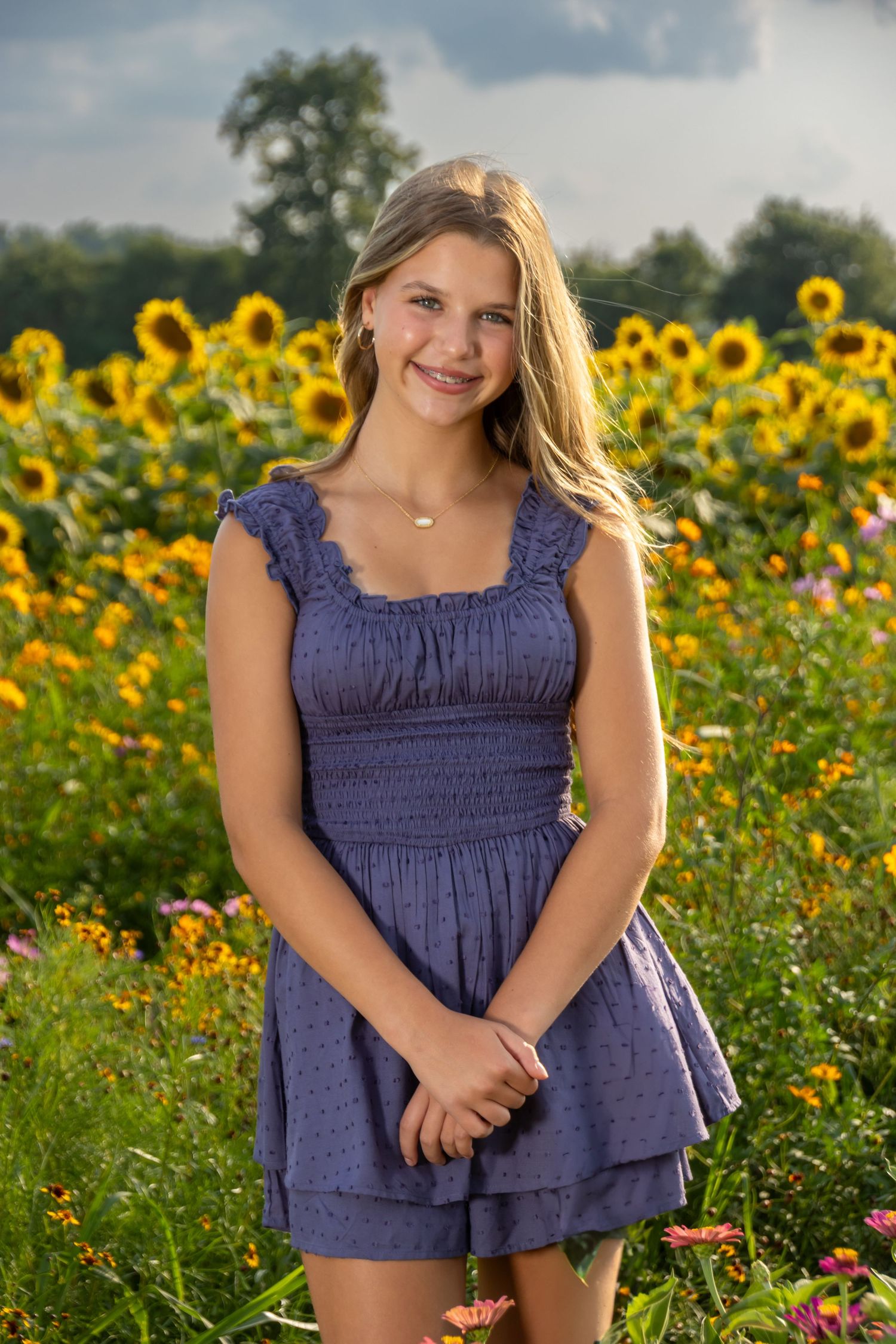 Smiling teenage girl in blue dress standing in a vibrant sunflower field on a summer day. Salem and Gloucester County Children Portraits