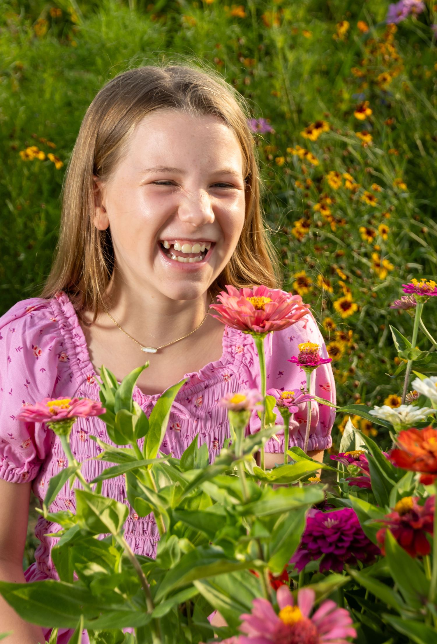 A laughing girl in a pink dress surrounded by colorful zinnia flowers in a sunny garden. Salem and Gloucester County Children Portraits