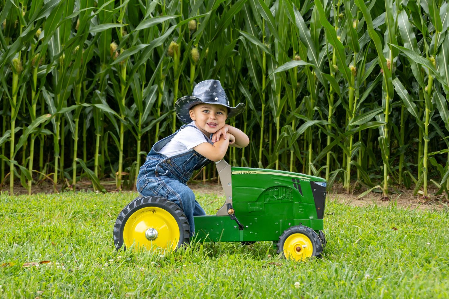 Young boy in overalls and cowboy hat riding a green John Deere toy tractor in front of a cornfield. Salem and Gloucester County portraits