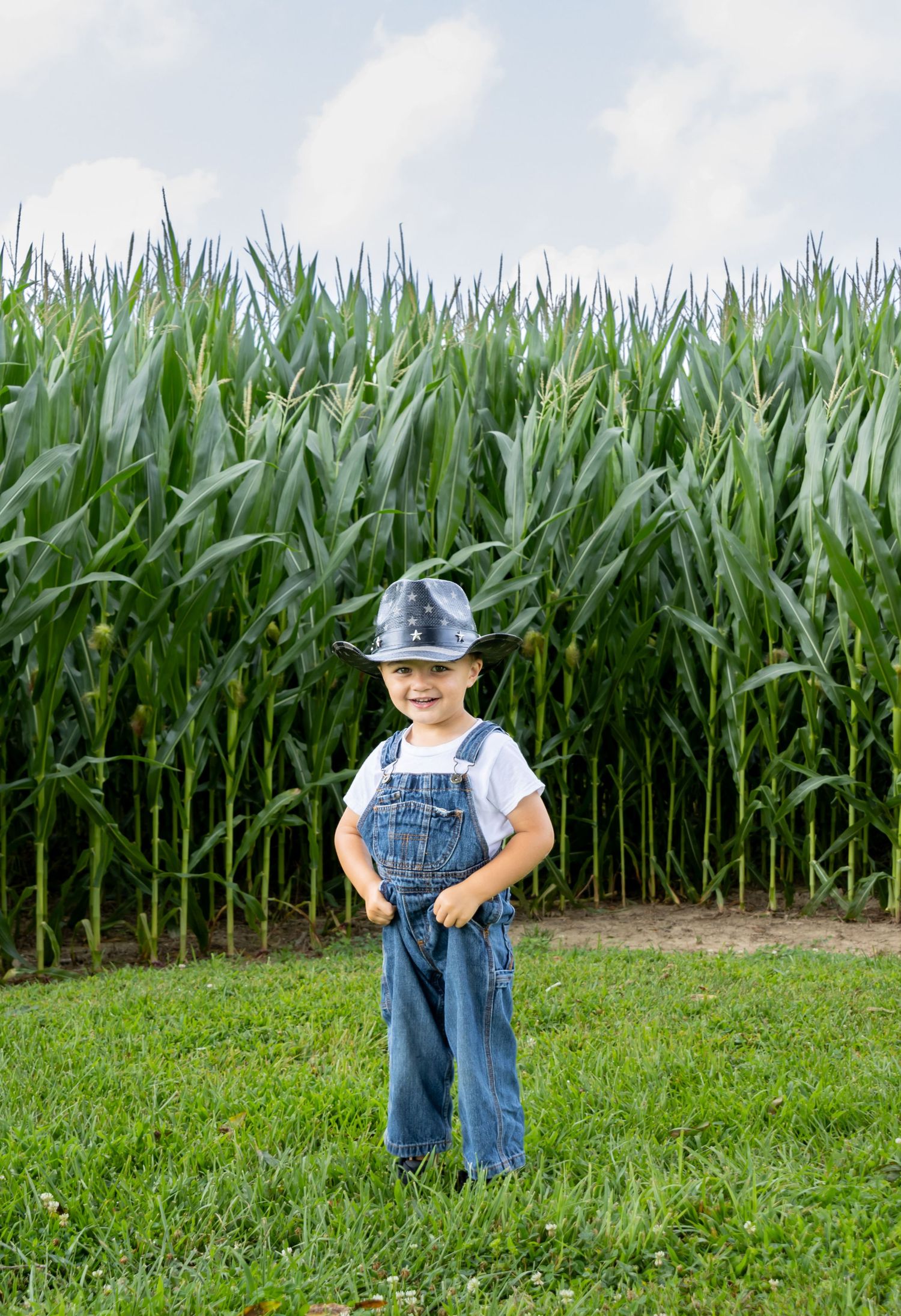 A young boy in denim overalls and a cowboy hat smiles while standing in front of a tall green cornfield. Salem & Gloucester County portraits