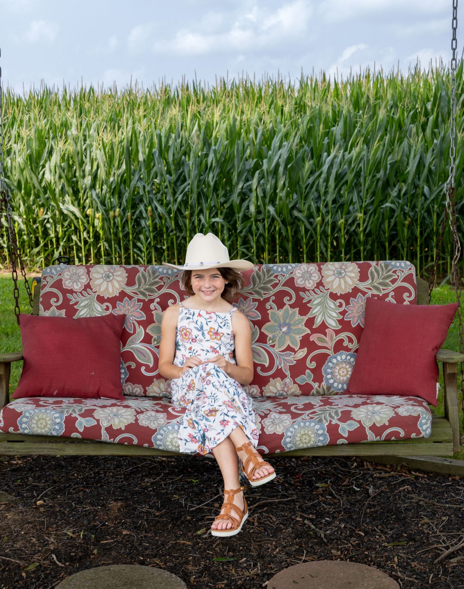 Young girl in floral dress and white cowboy hat sits on a swing bench near a cornfield. Salem and Gloucester County Children Portraits