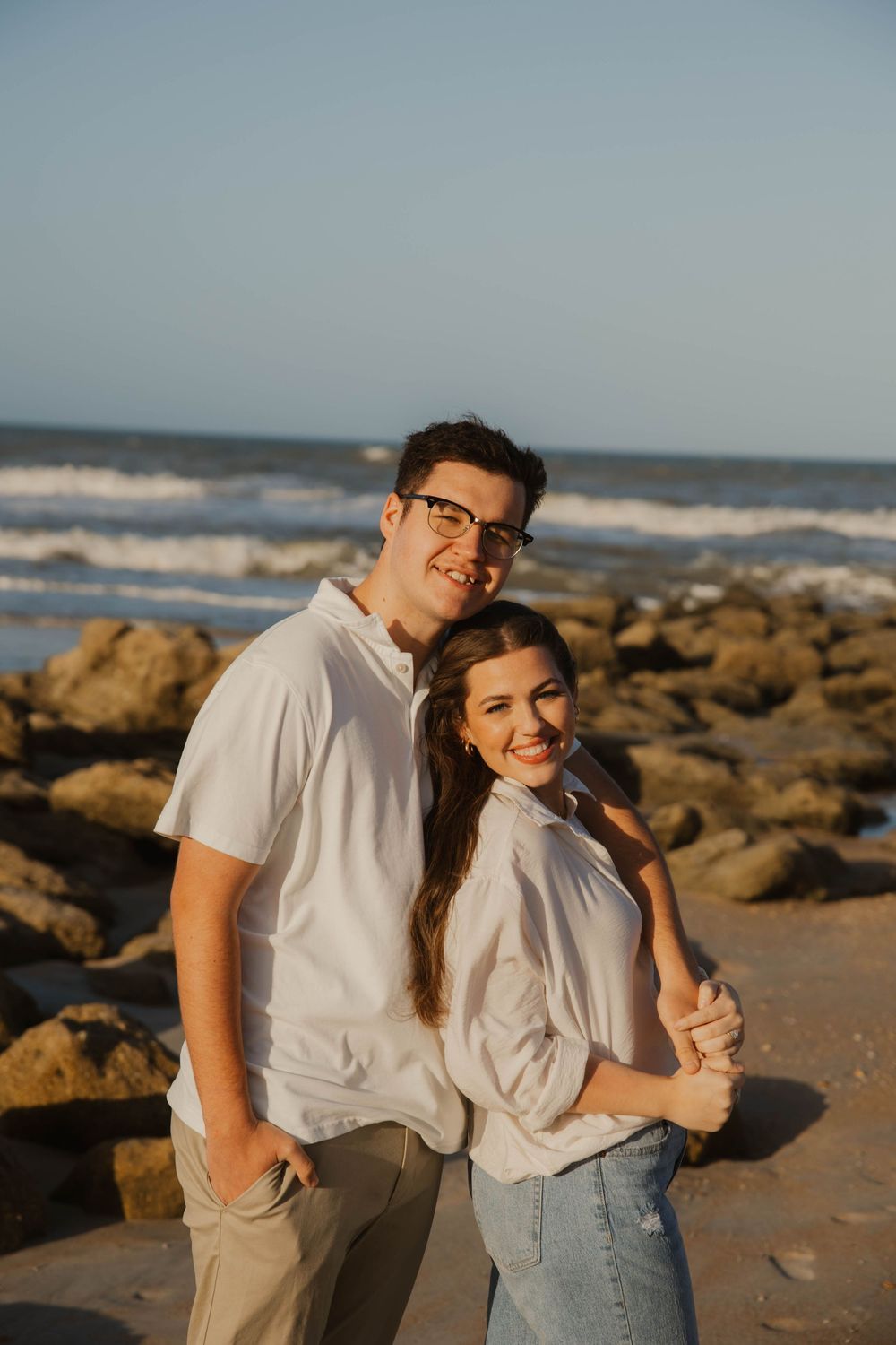 Couple in white shirts poses together on rocky beach at sunset.