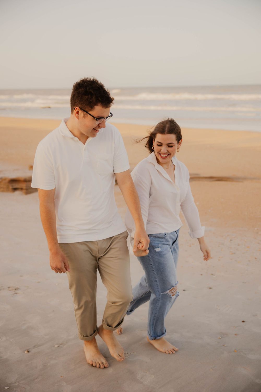 Barefoot couple walking and laughing together on sandy beach at sunset.