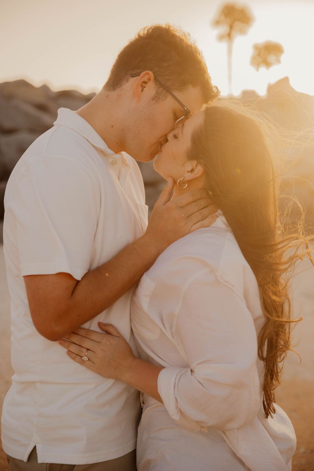 Loving couple embracing in warm sunset light on beach wearing white shirts.