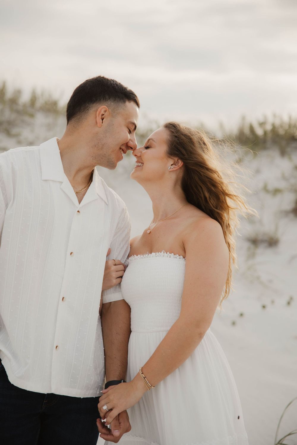 Romantic beach photoshoot showing couple embracing on white sand dunes at sunset.