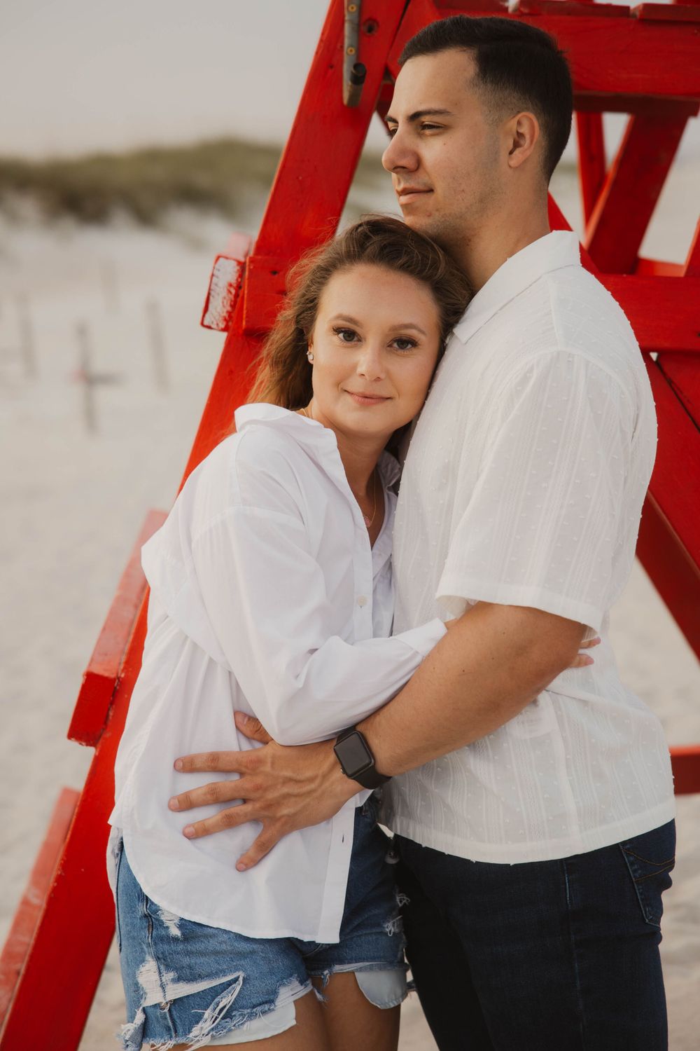 Couple in white shirts and denim embracing near red beach structure.