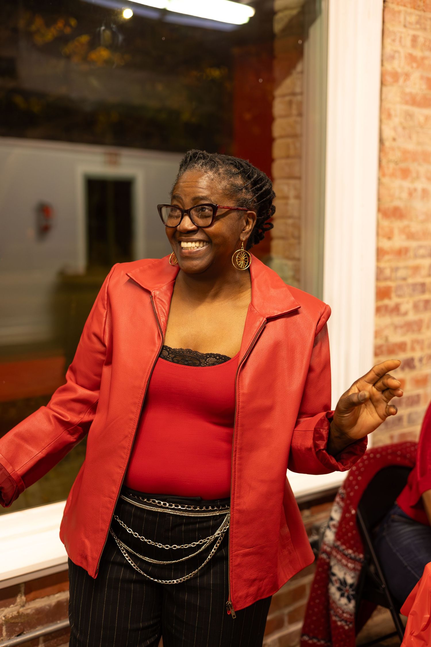 Person in red jacket and black striped pants smiling against brick wall backdrop.