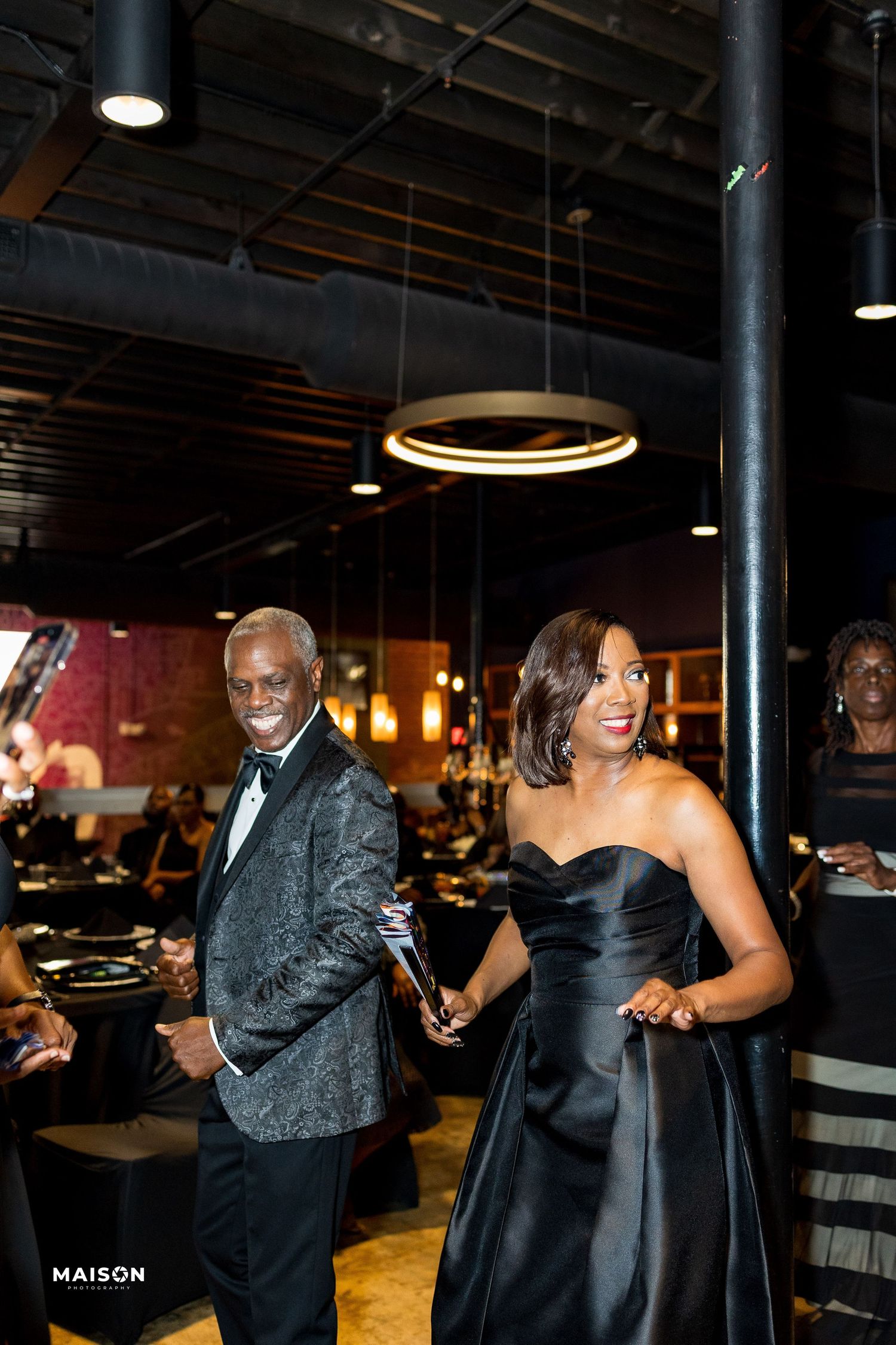Elegant guests in formal attire, including a silver patterned suit jacket and black strapless gown, at an evening event.