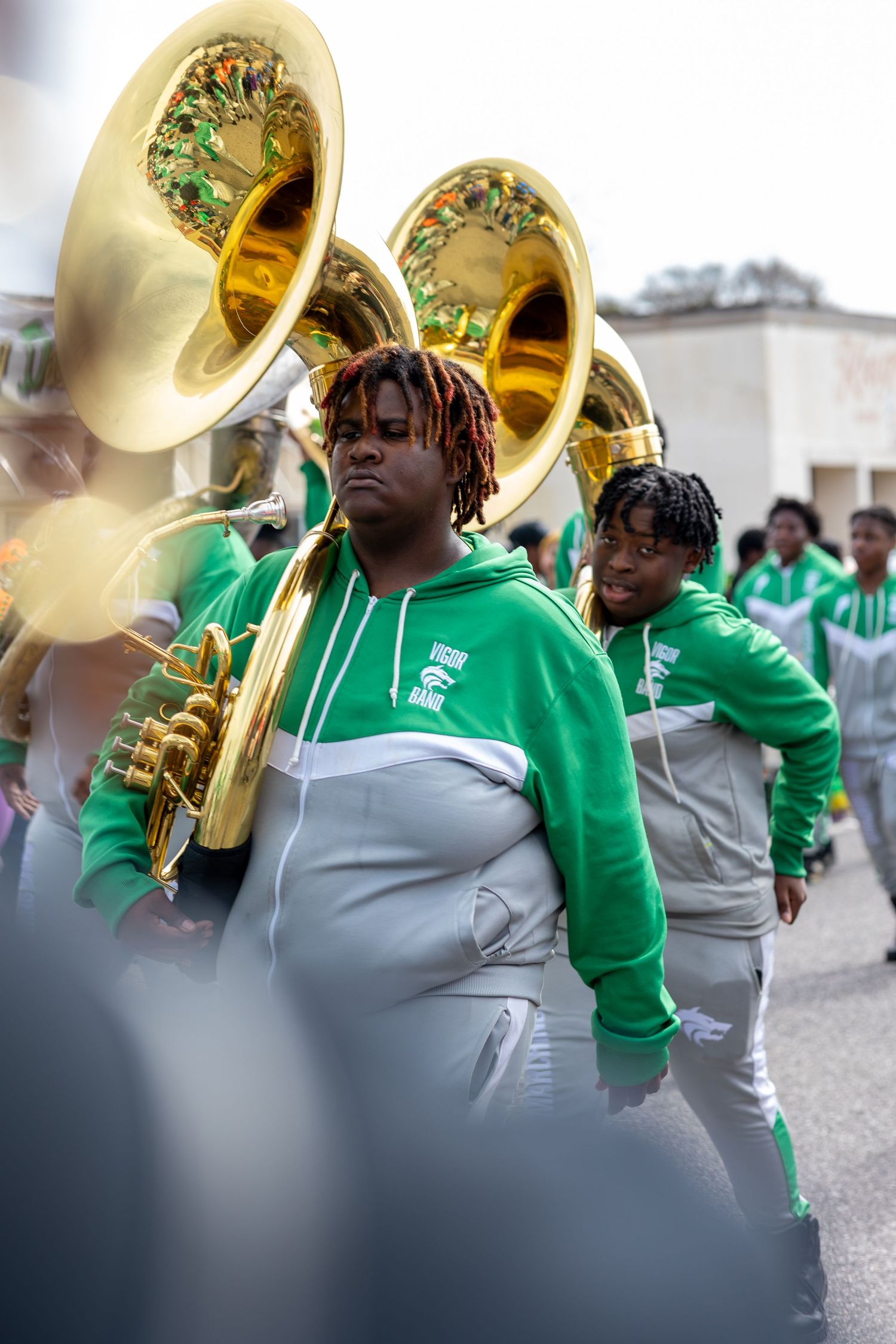Marching band members in green uniforms play brass instruments during a parade.