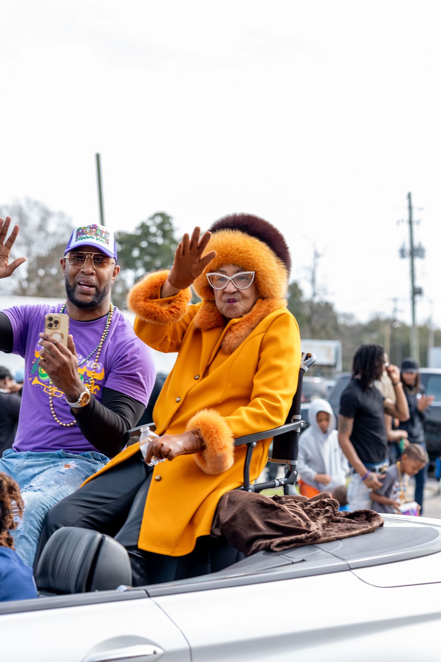 Person in bright orange coat waves from convertible during outdoor parade celebration.