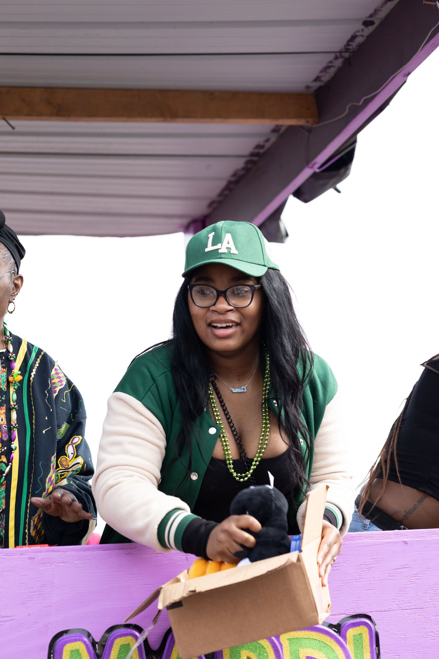 Participant wearing green baseball cap and varsity jacket at Mardi Gras celebration.