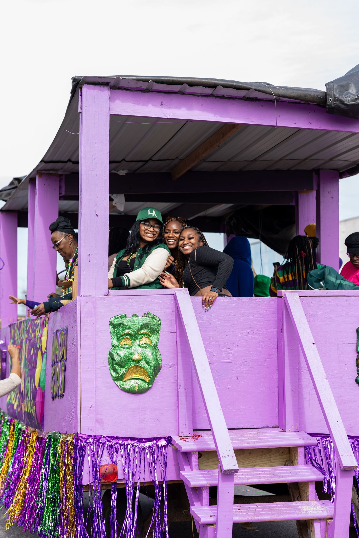 Revelers enjoy festivities from purple Mardi Gras parade float with green mask decoration.