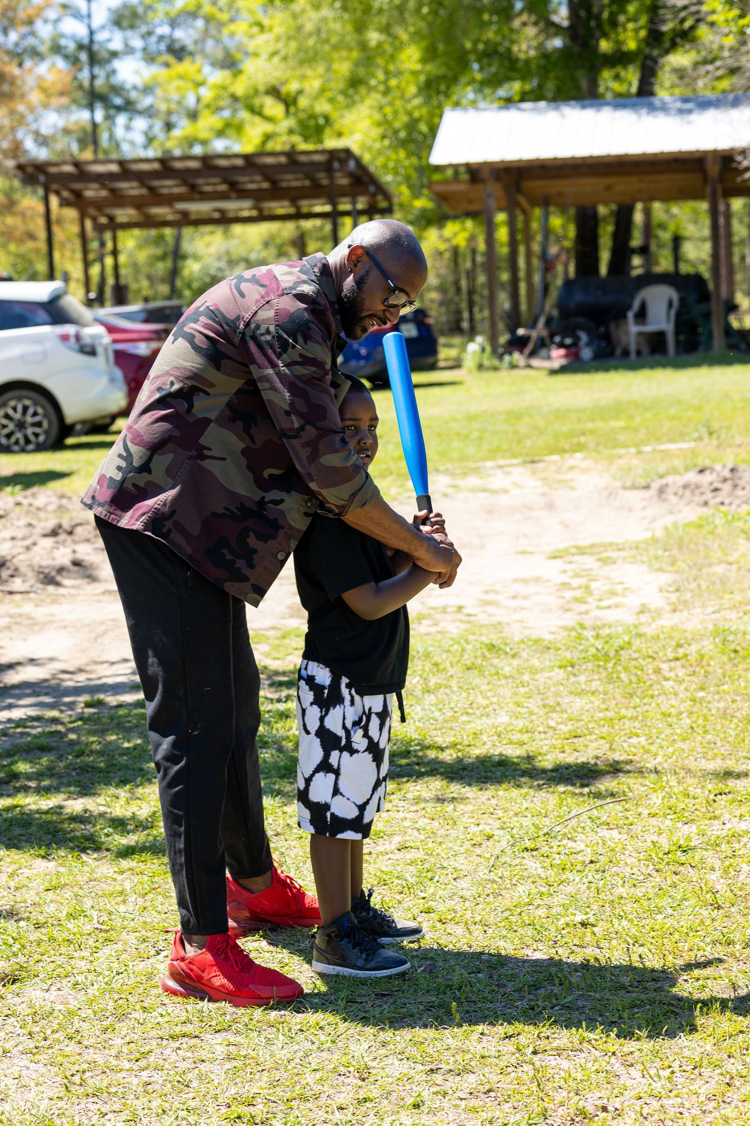 An adult helps adjust a child's clothing outdoors on a sunny day near parked cars.