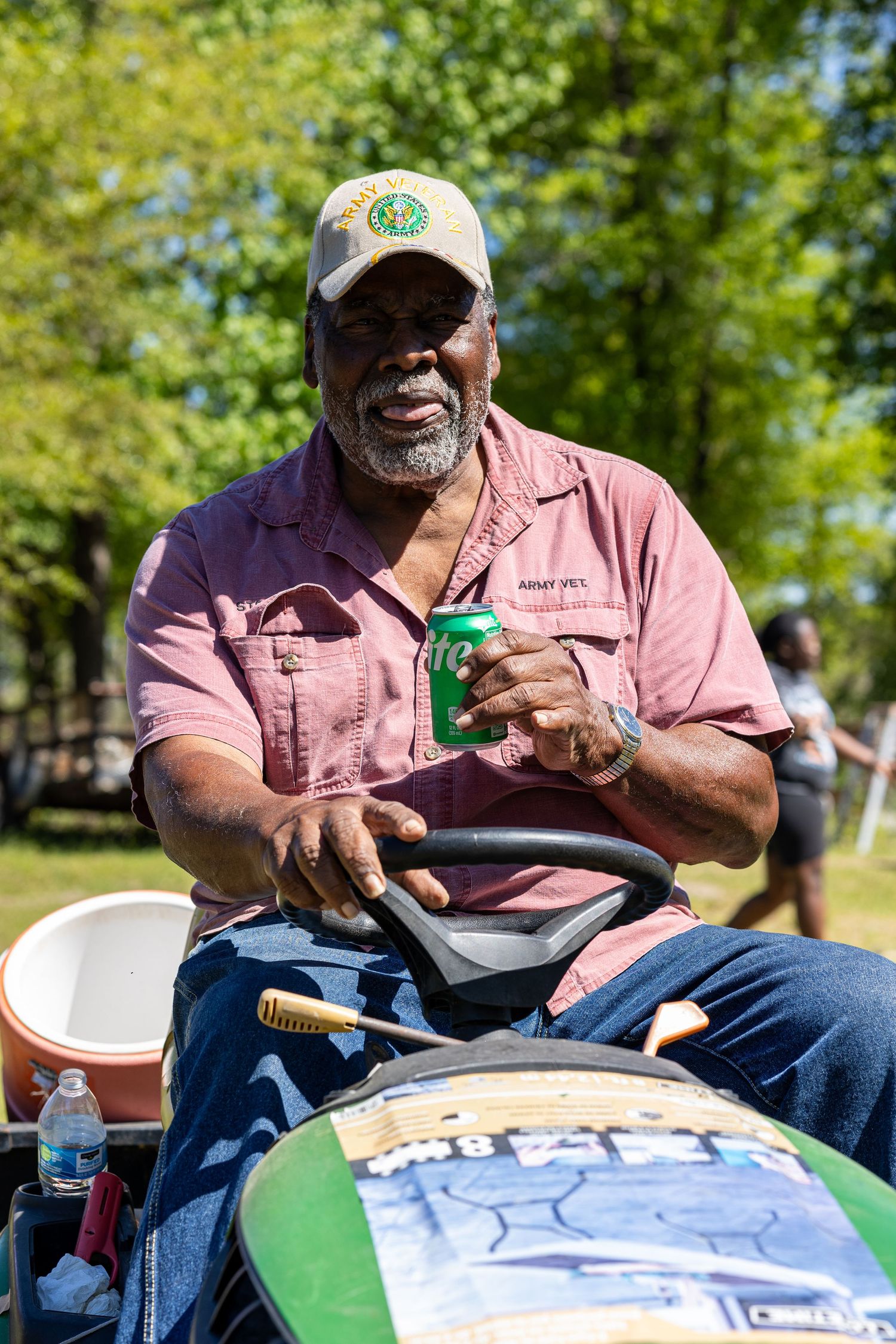 An older person in pink shirt and cap sits outdoors enjoying a beverage at a park gathering.