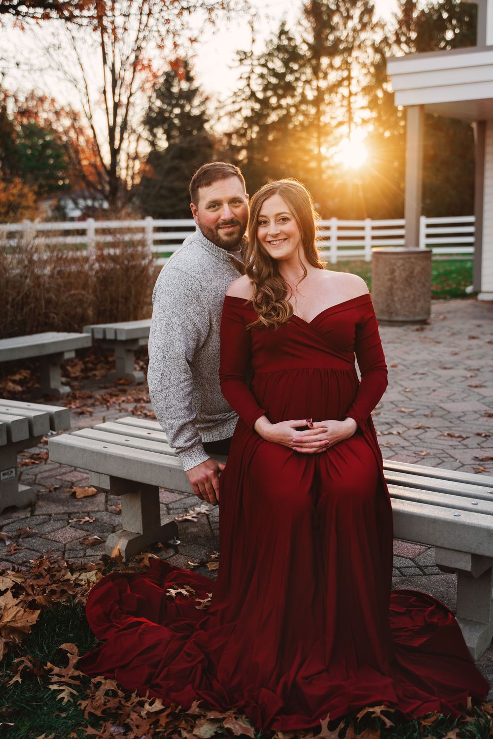 Pregnant couple posing at sunset in park, woman in elegant red maternity gown.