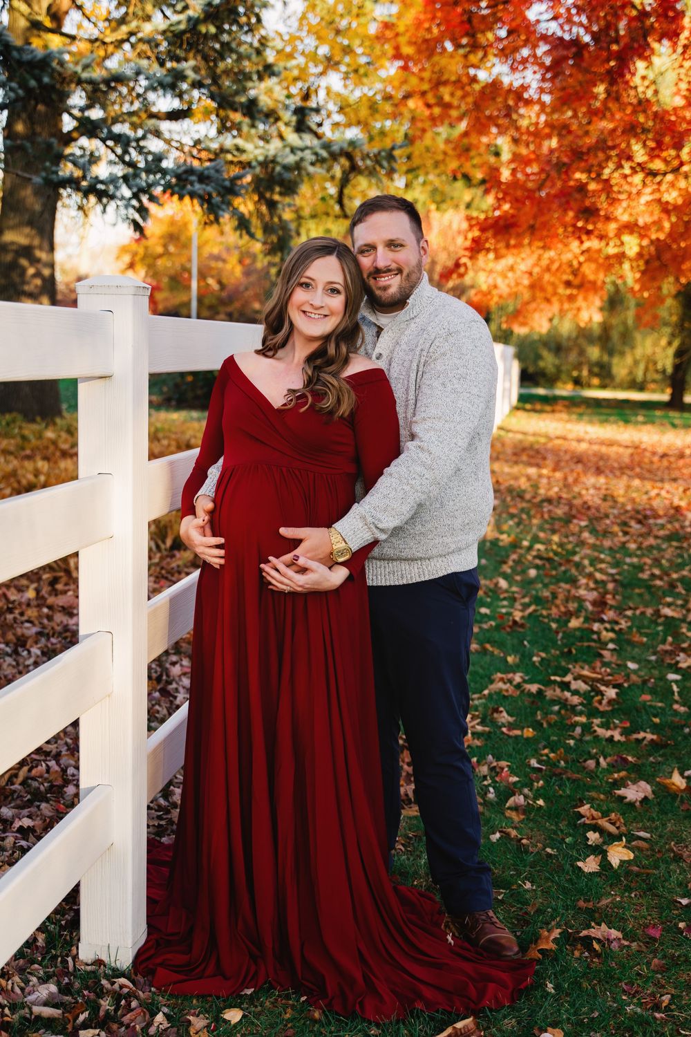 Smiling couple posing by white fence in fall foliage, woman in flowing red maternity dress.