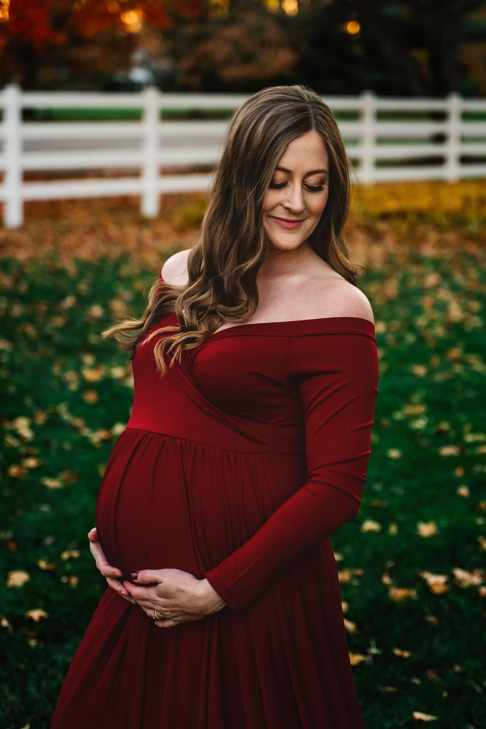 Pregnant woman in off-shoulder red maternity dress cradling baby bump outdoors in fall.