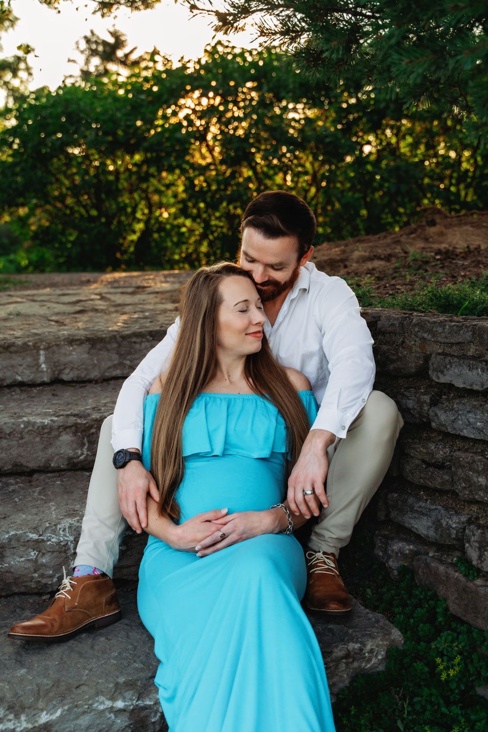 Expecting couple sitting together on stone steps at sunset, man tenderly embracing pregnant partner.