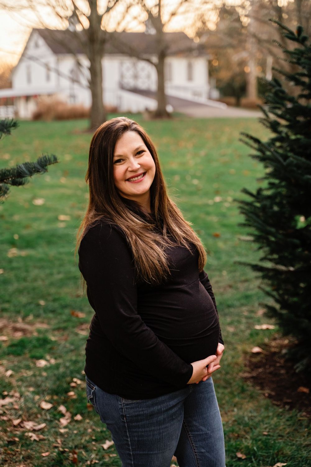 Smiling pregnant woman in black top and jeans cradling her baby bump near evergreen trees.