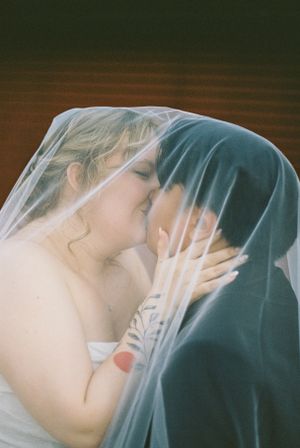 Romantic wedding kiss under a shared veil in soft lighting.