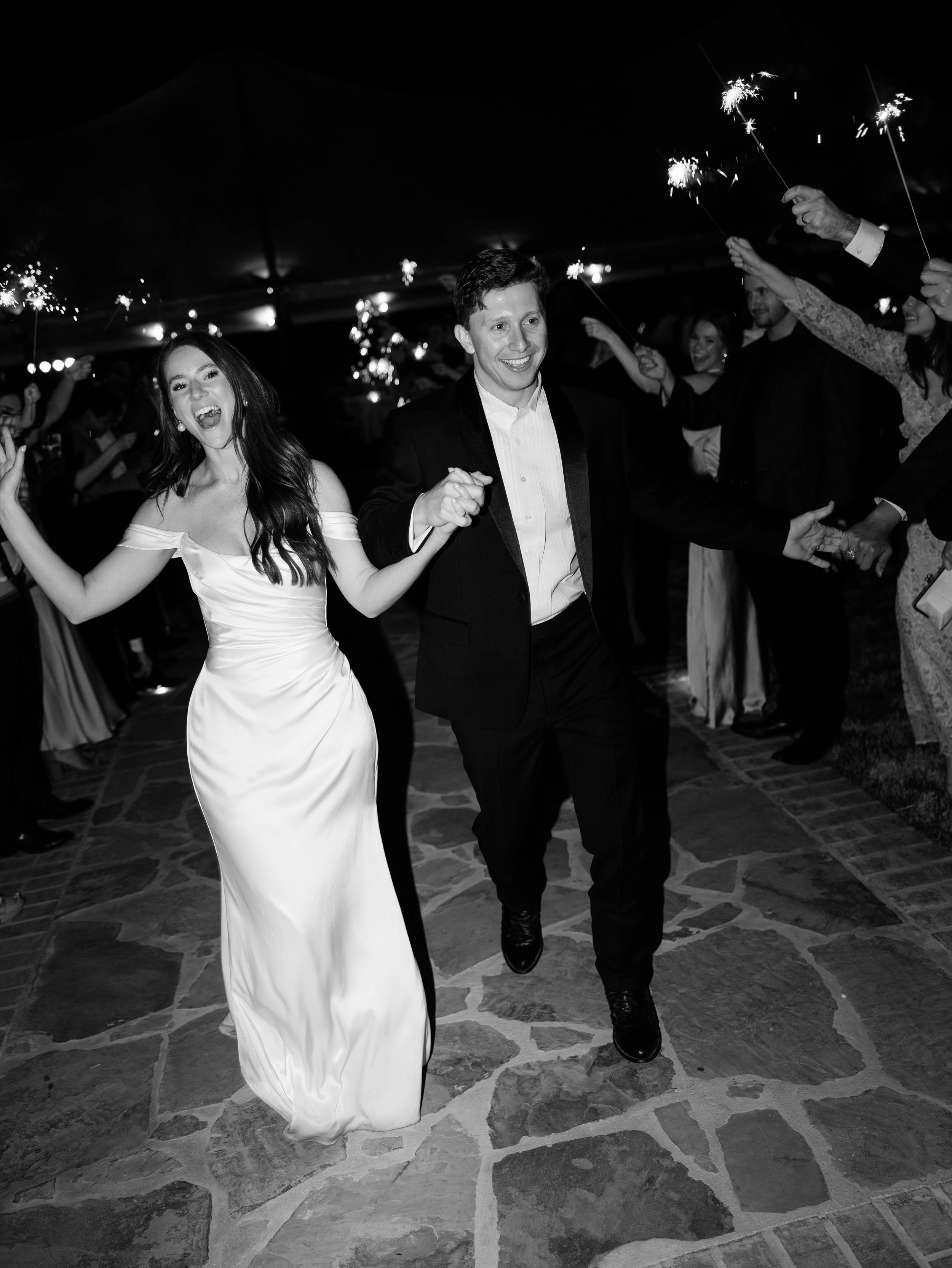 Black and white wedding photo of newlyweds joyfully running past sparklers held by guests on a stone paved path.