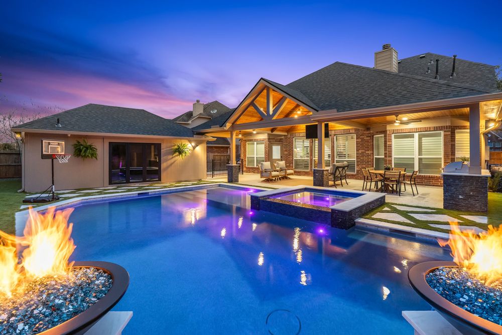 Luxury backyard pool with fire features, spa, and covered patio glowing under a twilight sky.