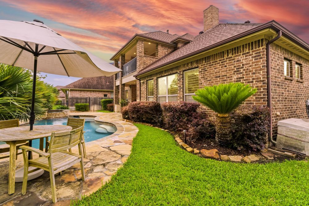 Brick home backyard with pool, stone patio, umbrella table, lush lawn, and tropical plants under a vibrant sunset sky.