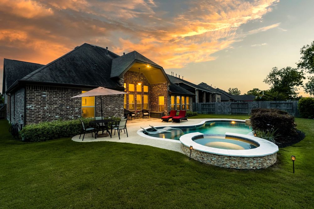Luxurious stone house with illuminated patio and curved swimming pool at sunset featuring dramatic orange sky.