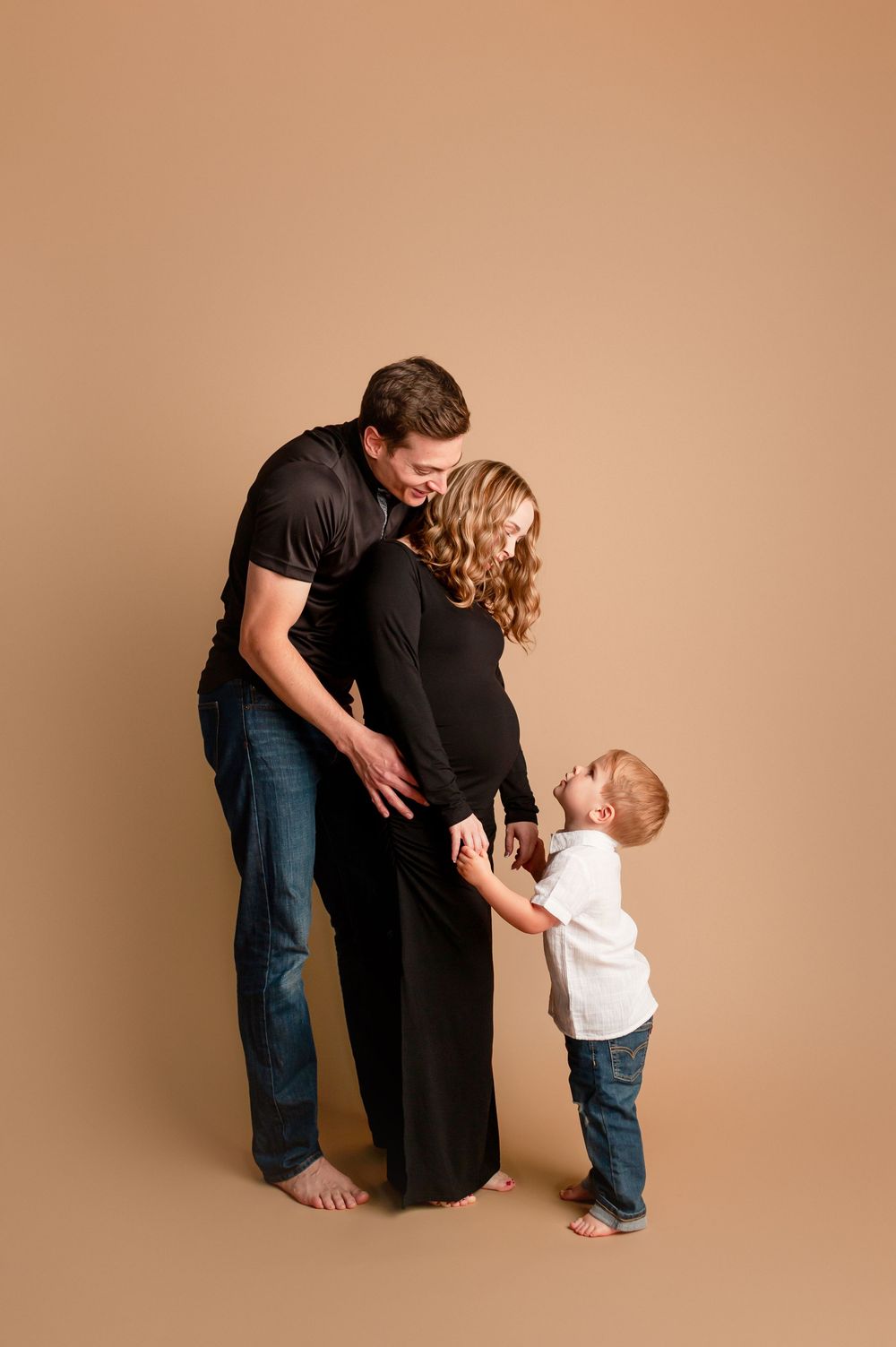 Pregnant woman in black dress with husband and toddler son looking down at her baby bump in a tan studio.