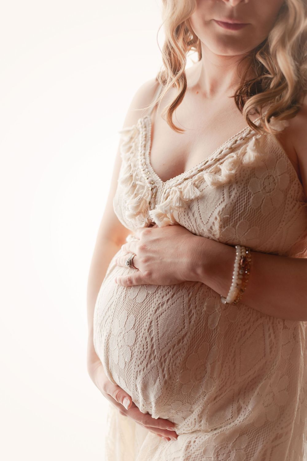 Close-up of a pregnant woman in a cream lace dress gently cradling her baby bump with pearl bracelet detail.