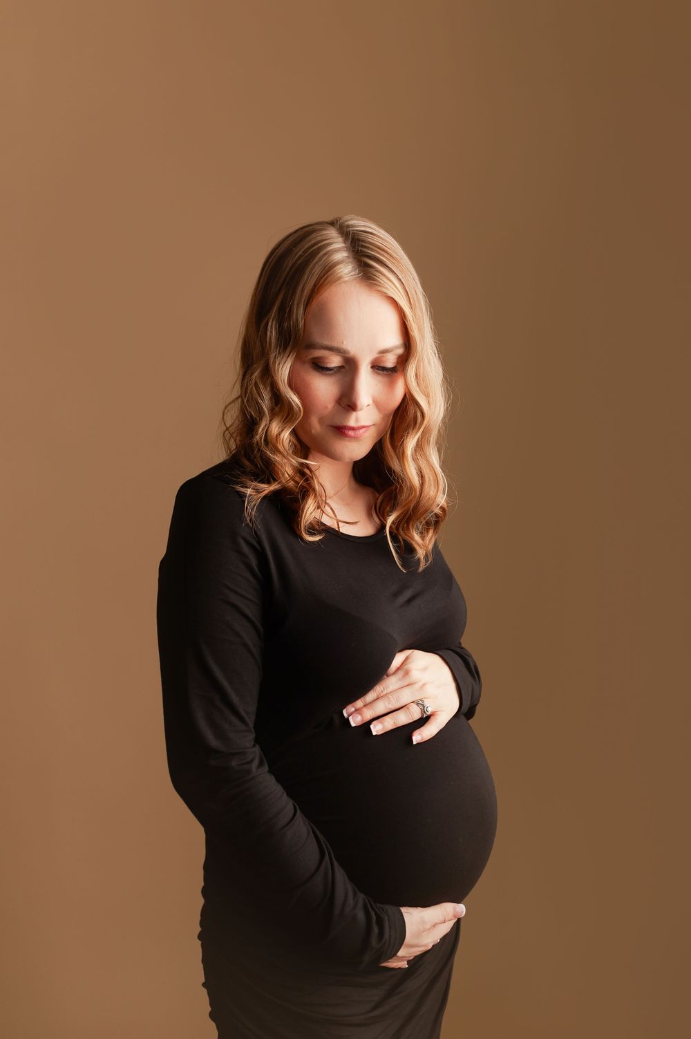 Pregnant woman in a black long-sleeve dress cradling her baby bump against a warm tan studio background.