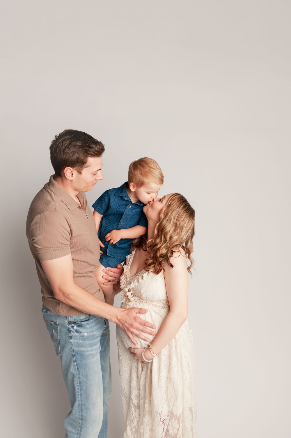 Expecting parents in a light studio, father holding toddler son who kisses pregnant mother in a white lace dress.