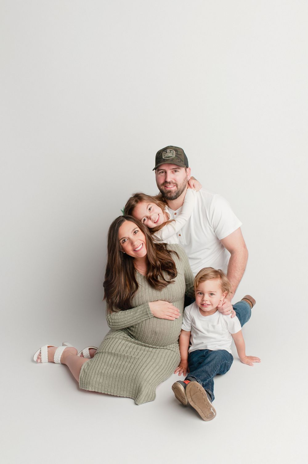 Happy family maternity photo with pregnant mom in green dress, dad in white shirt and cap, two young children.
