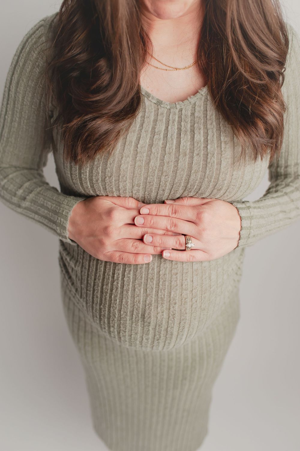Pregnant woman in sage green ribbed knit dress cradling baby bump with both hands, wearing engagement ring.