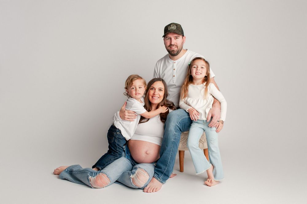 Family maternity portrait with pregnant mom, dad, and two kids sitting together on white studio background.