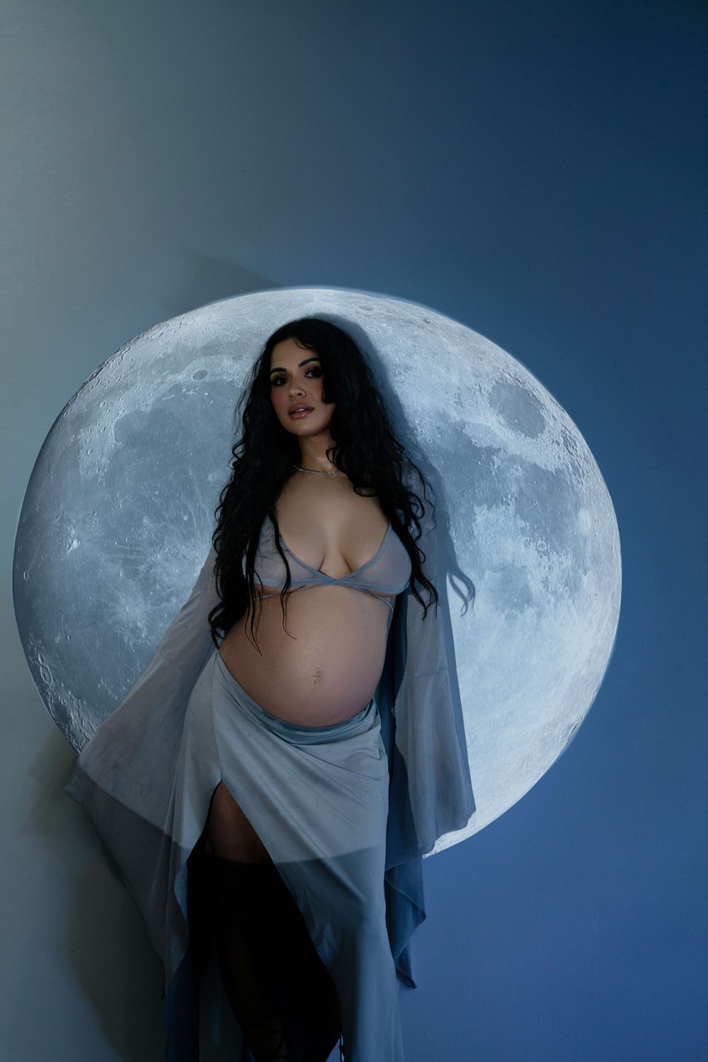 A maternity portrait against a large full moon backdrop with ethereal lighting and flowing fabric.