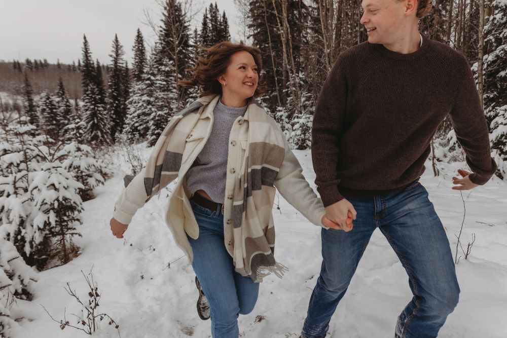 A couple running through a snowy winter forest holding hands.