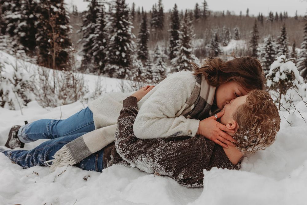 Couple lying romantically in the snow surrounded by snow-covered evergreen trees in a winter forest setting.