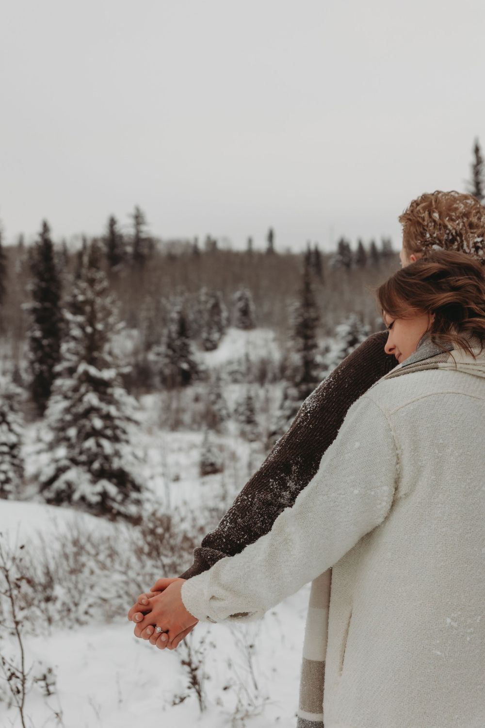 A pair stands together in a snowy winter landscape with snow-covered pine trees in the background.