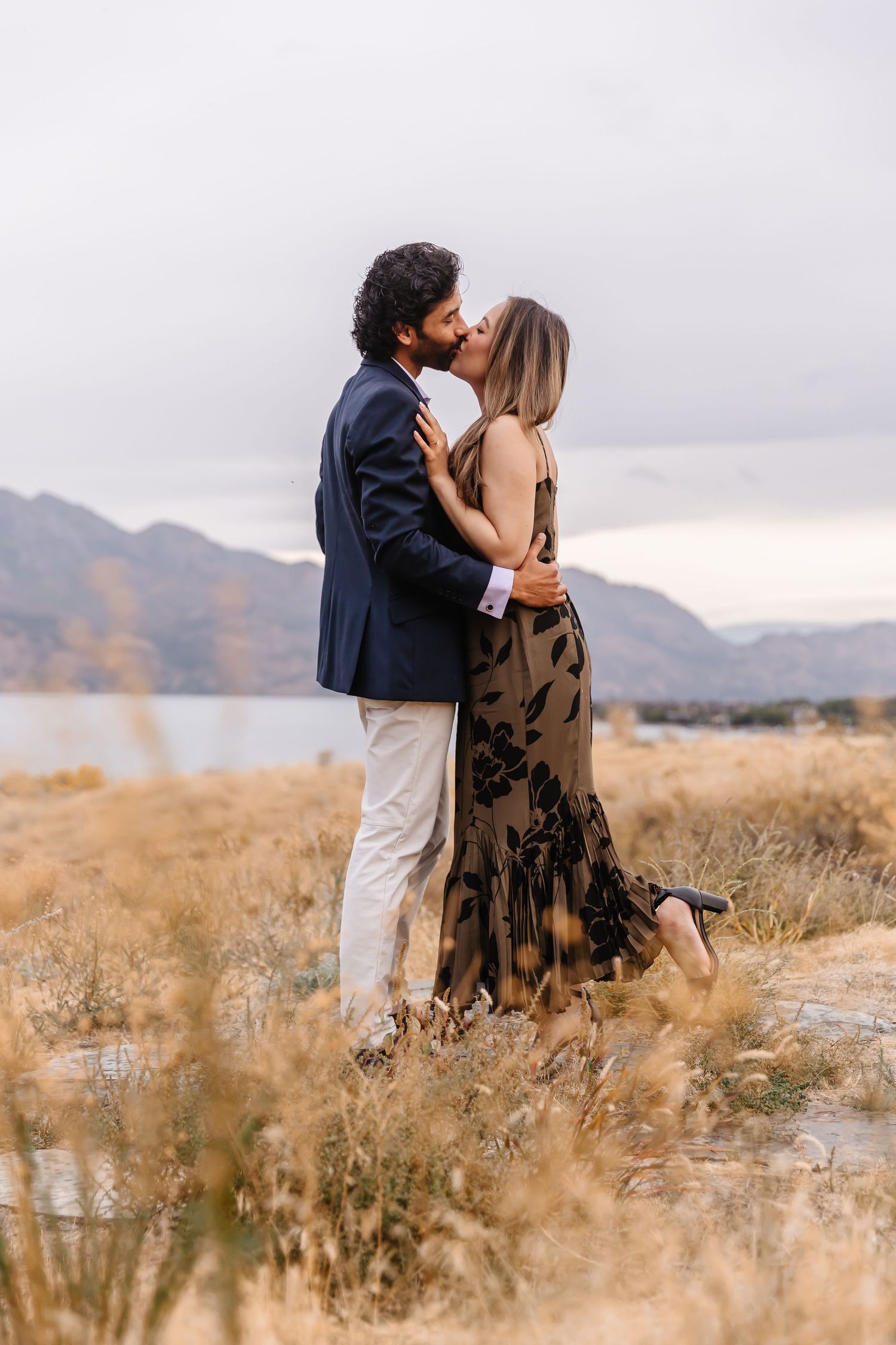 Couple sharing tender moment in tall grass with mountains and overcast sky.