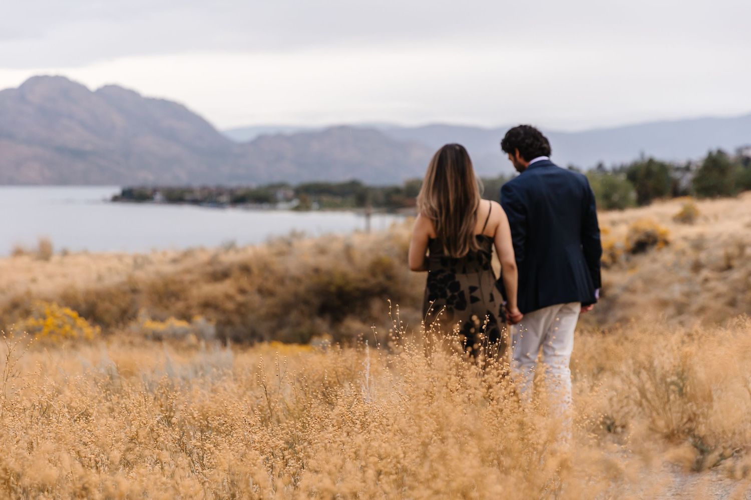 Couple walking through golden grass field with mountains visible in distance.