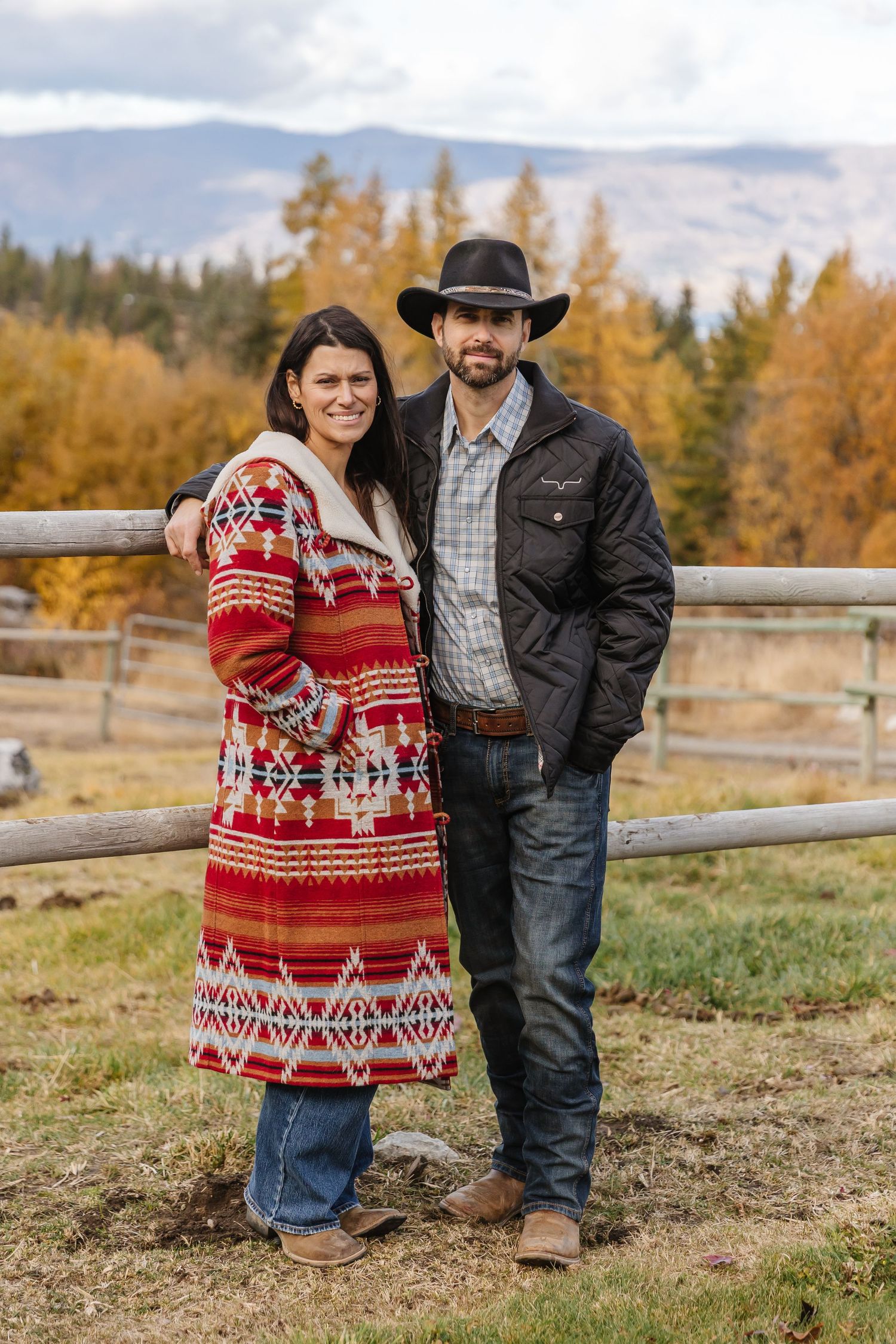 A couple stands together in autumn scenery with mountains and fall foliage, wearing western attire and a festive sweater.