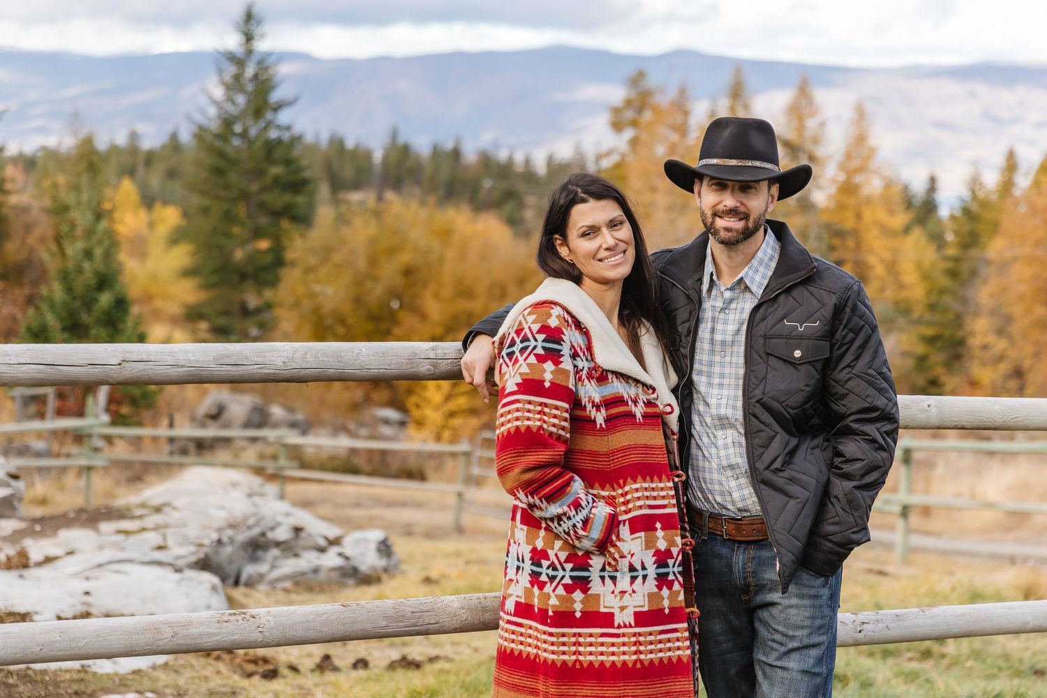 Couple in winter attire embracing outdoors during autumn with golden trees and wooden fencing in the background.