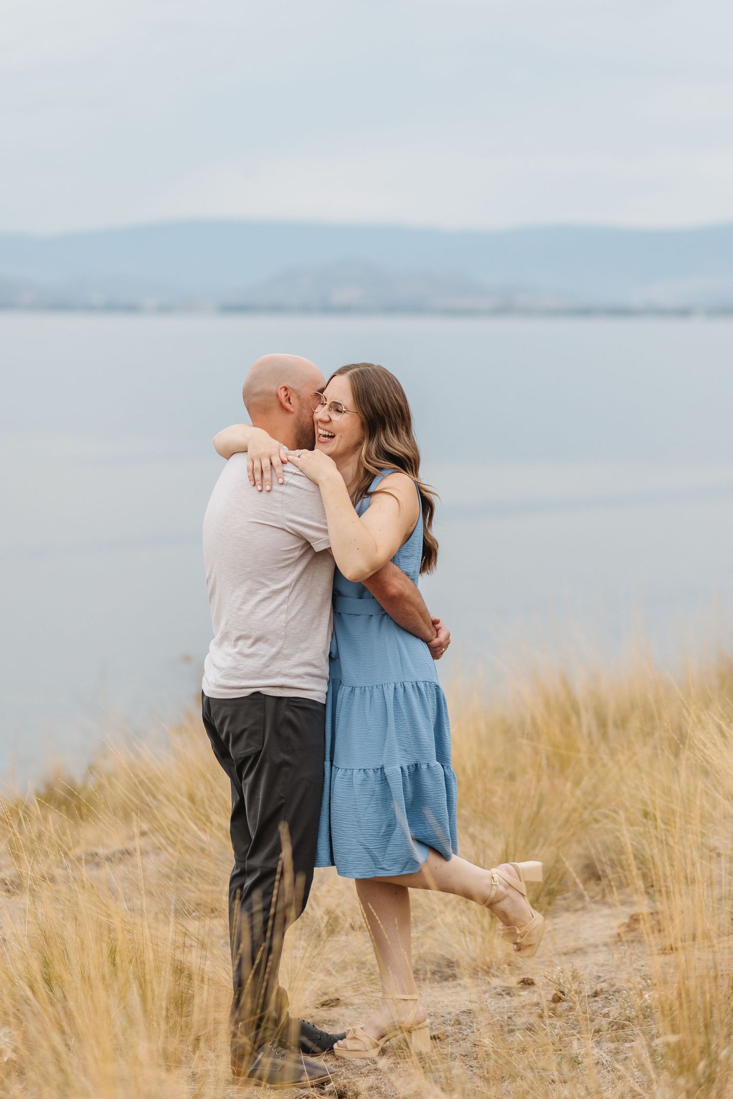 A couple embraces romantically on a beach with golden grass and calm waters in the background.