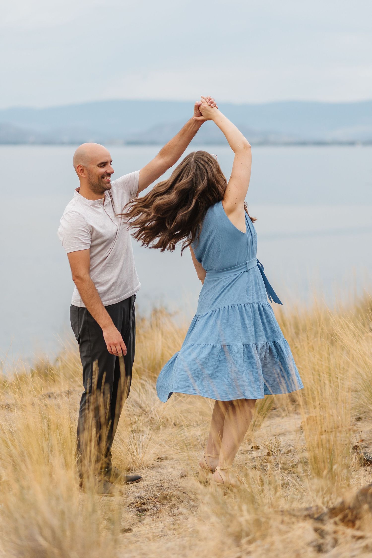 A couple dances together in tall grass by the ocean, with the woman wearing a flowing blue dress and spinning gracefully.