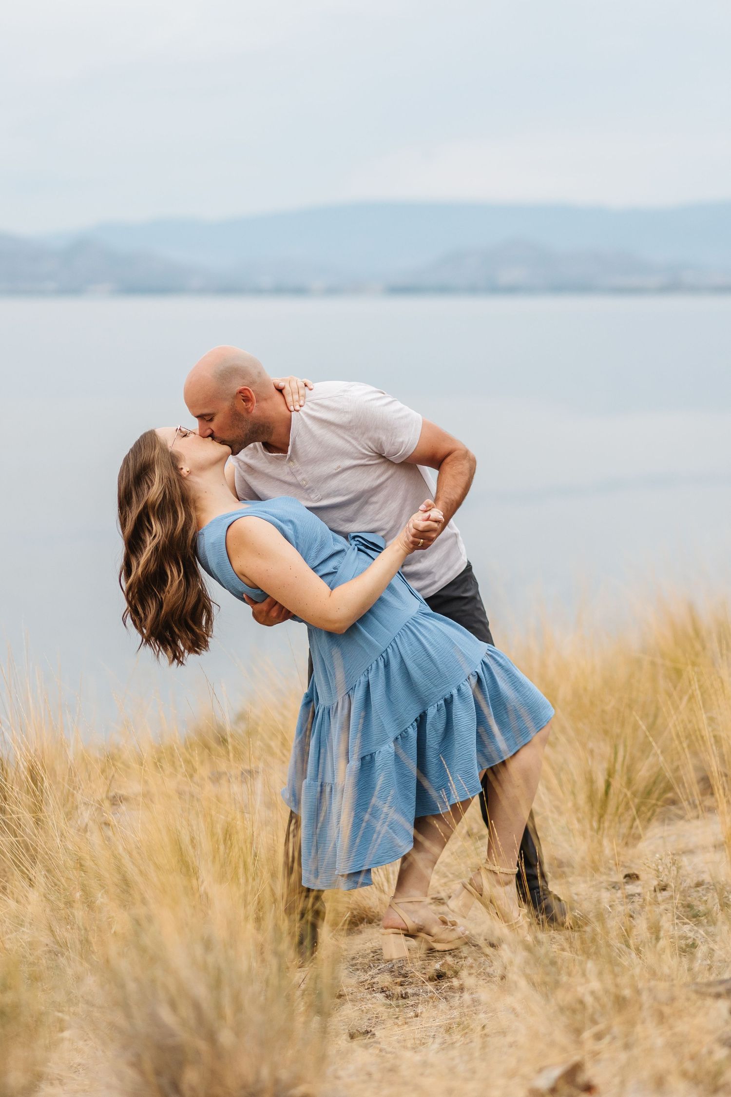 A romantic couple dances together in tall grass by a misty lake during sunset.