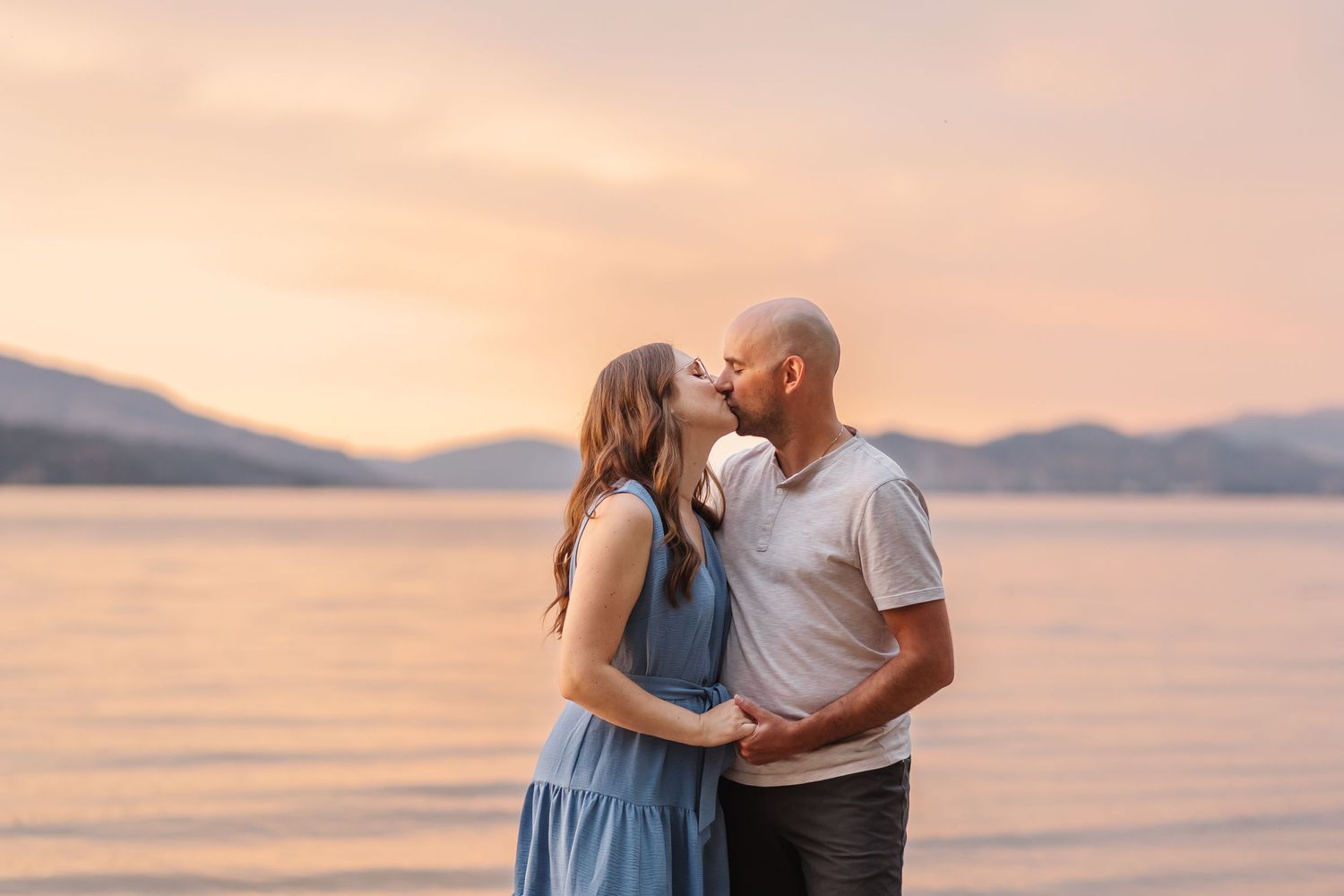 A couple shares an intimate moment at sunset by the ocean with mountains in the background.