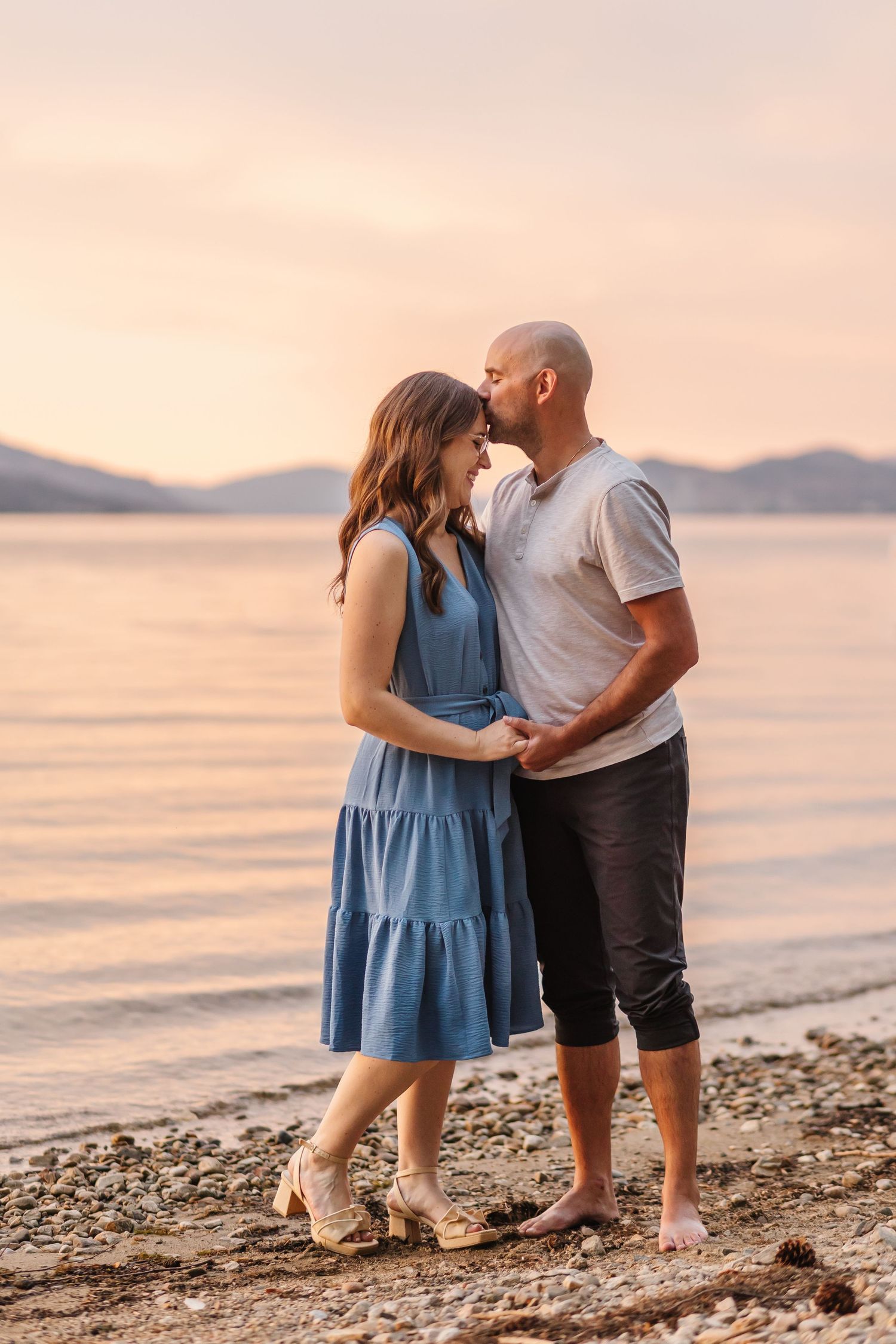A couple embraces on a beach at sunset with mountains and calm water in the background.