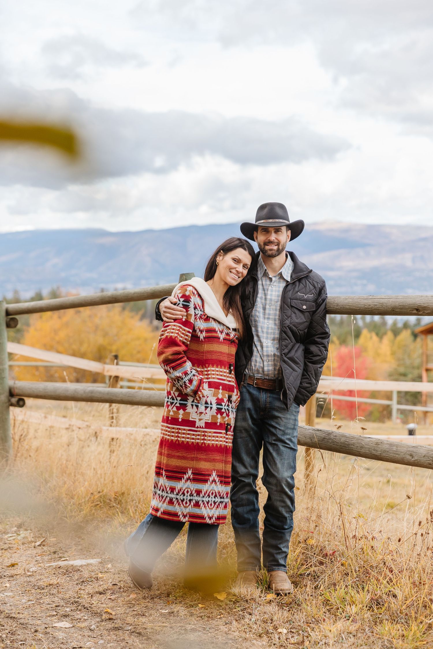 A couple poses by a rustic wooden fence with mountain views and fall colors in the background.