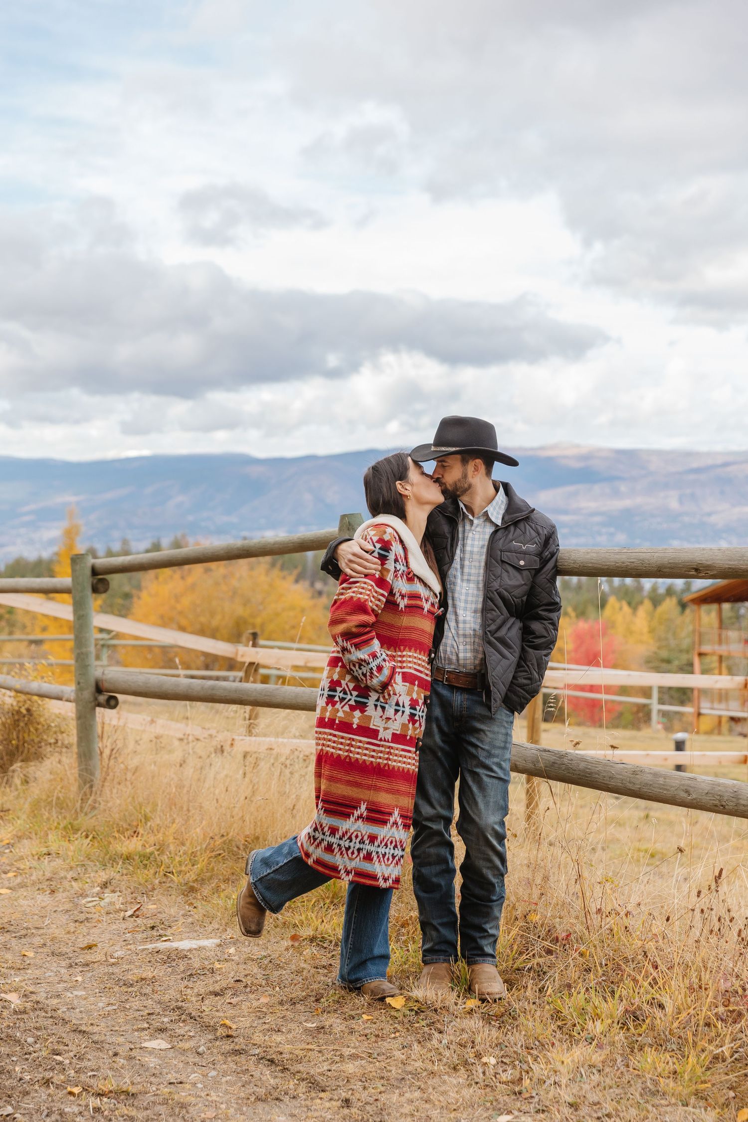 Couple in western attire sharing romantic moments by rustic wooden fence during autumn in mountain landscape.